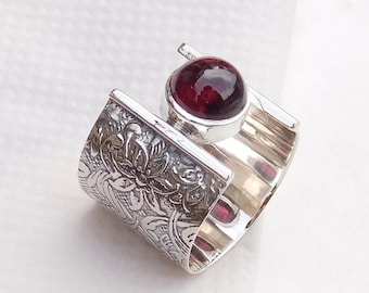 May include: A silver ring with a dark red gemstone set in a square bezel. The band of the ring is textured with a floral design.