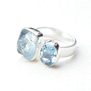 Puede incluir: Anillo de plata con dos piedras preciosas azul claro. Una es de forma rugosa e irregular, la otra de talla ovalada. El anillo tiene un acabado pulido y brillante. Las piedras son de color azul pálido.