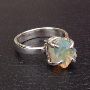 Könnte beinhalten: Ein silberner Ring mit einem rohen, hellblauen Opal-Edelstein, der in einer Krappenfassung sitzt. Der Opal hat eine natürliche, unregelmäßige Form und zeigt Farbschimmer.