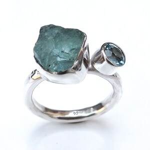 K&ouml;nnte beinhalten: Ein silberner Ring mit zwei Aquamarin-Steinen. Der gr&ouml;&szlig;ere Stein ist ein roher, nat&uuml;rlicher Aquamarin, w&auml;hrend der kleinere Stein ein runder, facettierter Aquamarin ist. Der Ring ist innen mit "925" gekennzeichnet.
