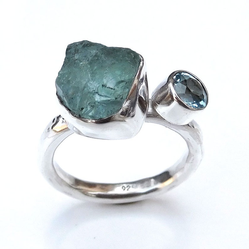 K&ouml;nnte beinhalten: Ein silberner Ring mit zwei Aquamarin-Steinen. Der gr&ouml;&szlig;ere Stein ist ein roher, nat&uuml;rlicher Aquamarin, w&auml;hrend der kleinere Stein ein runder, facettierter Aquamarin ist. Der Ring ist innen mit "925" gekennzeichnet.