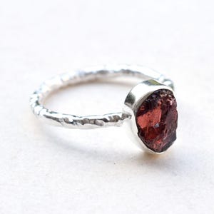 Puede incluir: Un anillo de plata con una banda rugosa y una gema grande, en bruto, de color rojo oscuro. La gema, de forma ovalada, está engastada en un bisel de plata liso. El anillo se muestra sobre una superficie blanca.