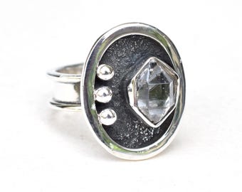 Può includere: Un anello d'argento con una pietra preziosa trasparente sfaccettata incastonata in una lunetta circolare testurizzata. Tre piccole perle d'argento sono visibili sul lato dell'anello.