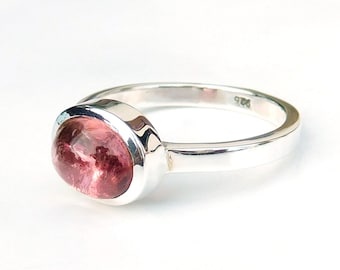 May include: A sterling silver ring featuring a large, oval-shaped pink gemstone. The gemstone is set in a bezel setting, and the band is smooth and polished. The ring is displayed against a white background.