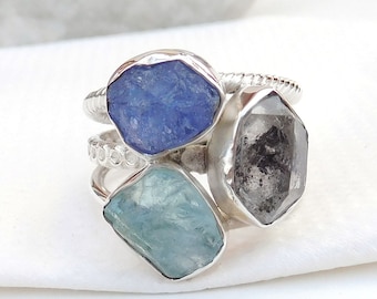 May include: A silver ring with three raw gemstones: a blue topaz, a clear quartz, and a light blue aquamarine.