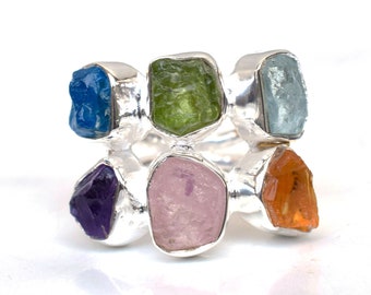 May include: A silver ring with six different colored gemstones. The gemstones are set in a cluster and are surrounded by silver. The gemstones are blue, green, light blue, pink, orange, and purple.