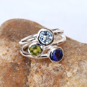 Könnte beinhalten: Ein silberner Ring mit drei runden Edelsteinen. Die Edelsteine sind blau, grün und lila.