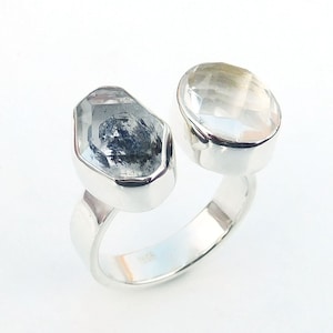 Könnte beinhalten: Ein silberner Ring mit zwei klaren Quarzsteinen. Die Steine sind in einer Zargenfassung gefasst und durch eine silberne Band verbunden.