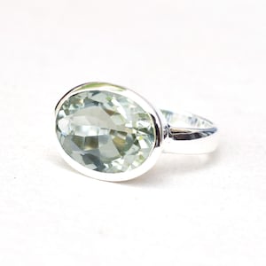Puede incluir: Un anillo de plata con una gran piedra preciosa ovalada de color verde pálido. La piedra preciosa está engastada en una banda de plata pulida. El anillo se muestra sobre un fondo blanco, resaltando la claridad y el color de la piedra.