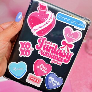 May include: A clear phone case with various colorful stickers. The stickers feature hearts, text, and illustrations. Some of the text includes "Just a Fantasy Romance Girlie", "Fake Dating", "Morally Grey", "Touch Her & Die Elixir", and "Careful, princess".