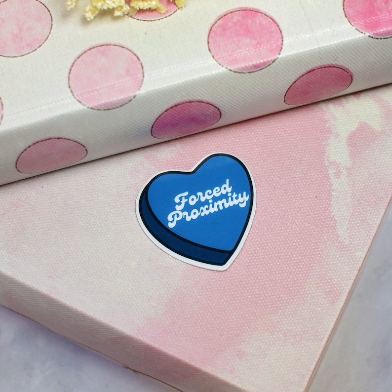 May include: A blue heart-shaped sticker with the text "Forced Proximity" in white lettering. The sticker is on a pink and white background.