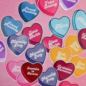 May include: A collection of heart-shaped stickers with various phrases, including "Forced Proximity", "Slow Burn", "Morally Grey", "One Bed", "Forbidden Romance", "Enemies to Lovers", "Second Chance", "Fake Dating", "Mine", and "Soul Mate".