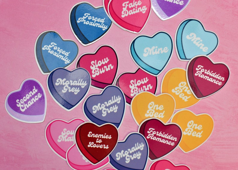 May include: A collection of heart-shaped stickers with various phrases, including "Forced Proximity", "Slow Burn", "Morally Grey", "One Bed", "Forbidden Romance", "Enemies to Lovers", "Second Chance", "Fake Dating", "Mine", and "Soul Mate".