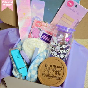 May include: A gift box filled with various items for book lovers, including bookmarks, a book club sticker, a glass water bottle with the text "Just One More Chapter", a coaster with the text "I Read Past My Bedtime", a blue and white checkered blanket, and a blue light.