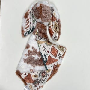 Puede incluir: Una manta para bebé y un juguete de orejas de conejo. La manta presenta un estampado de piel de vaca por un lado y un diseño desértico del suroeste por el otro. Las orejas de conejo son marrones y blancas, con un estampado desértico a juego.