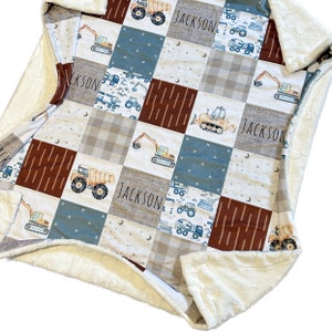May include: A patchwork baby blanket with a brown, blue, and white color scheme. The blanket features a variety of construction vehicles, including dump trucks, excavators, and bulldozers. The blanket is personalized with the name "JACKSON" in a handwritten font.
