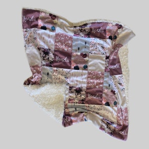 Puede incluir: Una manta suave y mullida para beb&eacute; con un dise&ntilde;o de patchwork que presenta patrones florales y el nombre "Henzli" repetido en toda la superficie. La manta tiene un respaldo de sherpa blanco.