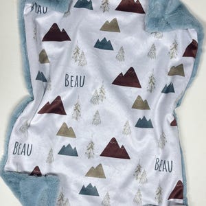 Puede incluir: Una manta para bebé con interior de satén blanco con un estampado de montañas y árboles en tonos marrón, verde y beige. La manta tiene un borde de piel sintética azul suave y el nombre "BEAU" está impreso.