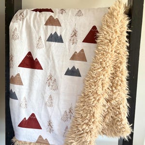 May include: A soft, white blanket with a mountain and tree pattern in shades of brown, burgundy, and gray. The blanket has a plush, tan faux fur trim. The blanket is draped over a black wooden ladder.