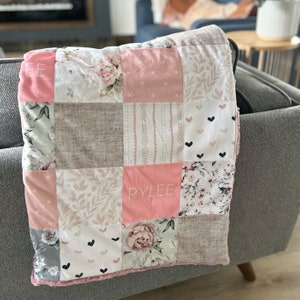 Peut inclure: Une couverture bébé en patchwork rose et gris avec le nom "RYLEE" brodé sur l'un des carrés.
