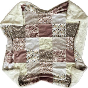 Peut inclure: Une couverture bébé en patchwork avec un dos blanc doux et moelleux. La couverture présente une variété de motifs floraux dans des tons de brun, beige et rose.