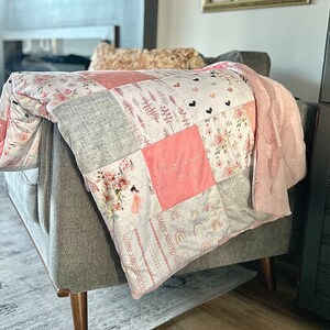 Peut inclure: Une courtepointe patchwork rose et grise avec un nom brodé personnalisé "Mckenna" dans un carré rose. La courtepointe est pliée et drapée sur un fauteuil gris.
