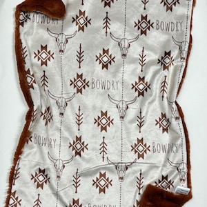 Puede incluir: Una manta para bebé blanca con un ribete de piel sintética marrón. La manta presenta un patrón repetido de calaveras de toro marrones, formas geométricas y la palabra "BOWDRY".