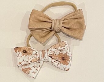 May include: Two hair bows, one brown and one with a floral pattern. The bows are tied with a knot in the center and are attached to elastic headbands.