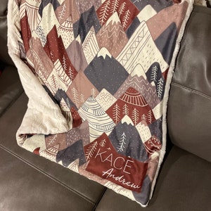 Puede incluir: Una manta suave y mullida con un diseño de montaña y bosque en tonos marrones, grises y rojos. La manta tiene un respaldo de piel sintética blanca y está personalizada con los nombres "Kace" y "Andrew".