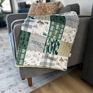 May include: A green, grey and white patchwork baby blanket with a sherpa backing. The blanket features a variety of woodland animal prints, including deer, bears and foxes. The blanket is personalised with the name "LINCOLN" in black text.