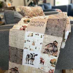 Puede incluir: Una manta de bebé de patchwork con temática de granja. La manta presenta un granero marrón, vacas, tractores y un molino de viento. La manta está personalizada con el nombre "HARTLEN WILLIAM".