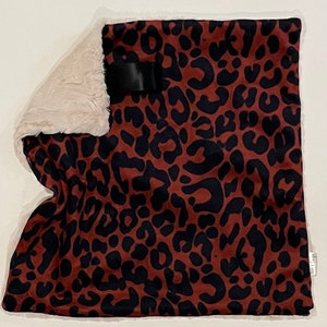 Puede incluir: Una almohada de color marrón y negro con estampado de leopardo y ribete de piel sintética blanca. La almohada tiene una cinta negra cosida en la parte delantera.