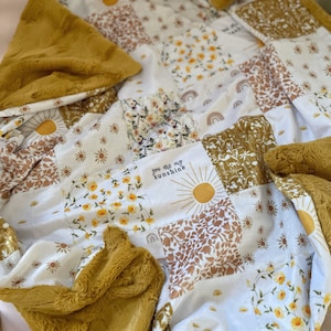 May include: A soft, plush blanket with a patchwork design featuring various floral and sun patterns. The blanket is made of a combination of faux fur and cotton fabric. The text "You are my sunshine" is embroidered on one of the patches.