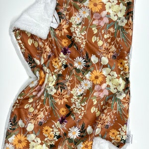 Puede incluir: Una manta de bebé floral marrón y blanca con un respaldo blanco suave y mullido. La manta presenta una variedad de flores en tonos de amarillo, blanco y rosa.