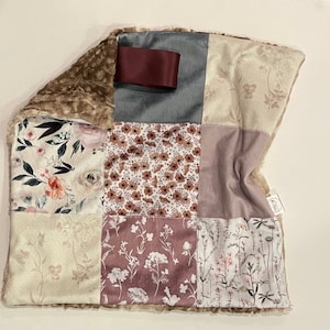 Peut inclure: Une couverture bébé en patchwork avec une bordure en fausse fourrure marron douce. La couverture est faite de différents tissus avec des motifs floraux dans des tons de rose, marron et blanc. Un tissu bleu avec un ruban bordeaux se trouve dans le coin supérieur gauche.