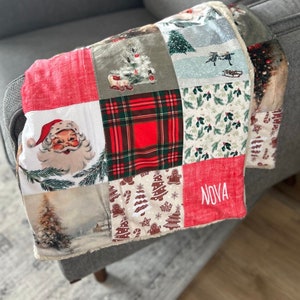 Peut inclure: Une courtepointe en patchwork avec un motif écossais rouge, vert et blanc, une image du Père Noël et un motif d'arbre de Noël. La courtepointe est personnalisée avec le nom "NOVA" en broderie blanche.