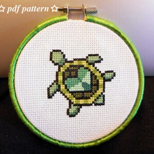 May include: A cross-stitch embroidery hoop featuring a turtle design. The turtle is stitched in shades of green, yellow, and brown against a white background. The hoop is trimmed with green satin ribbon. The text "pdf pattern" is at the top.