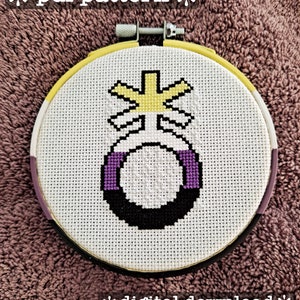 May include: A cross-stitch embroidery hoop with a white background and a purple, black, and yellow symbol in the center. The symbol is a circle with a cross on top and a purple band around the circle. The hoop is surrounded by a yellow, purple, and black border.