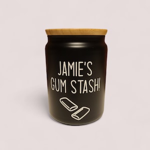 May include: Black ceramic jar with a wooden lid. The jar has white text that says "Jamie's Gum Stash!" and a graphic of two sticks of chewing gum.
