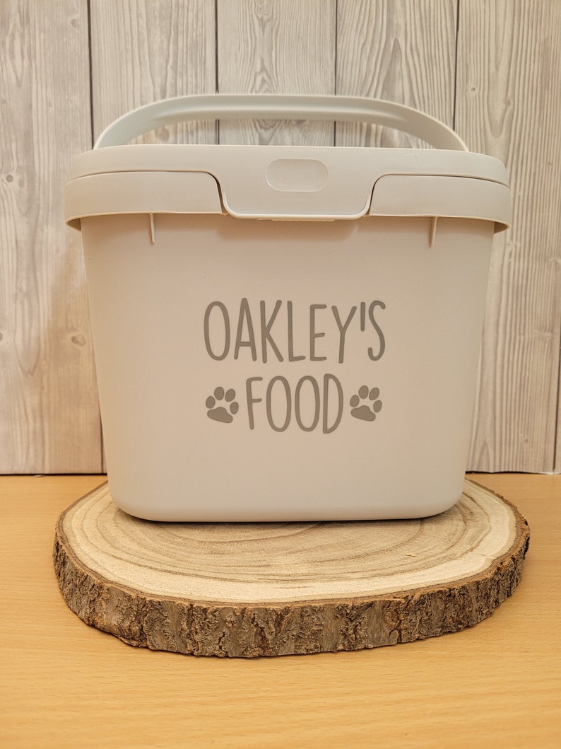 May include: A white plastic container with a handle and a lid. The container has the text "OAKLEY'S FOOD" with paw prints on either side of the word "FOOD". The container is sitting on a wooden slice.