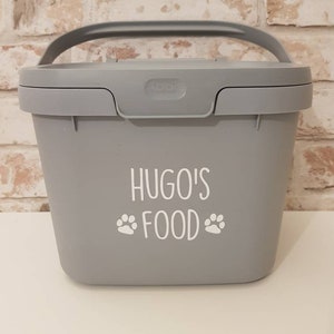 May include: A gray plastic container with a handle and a white label that says "Hugo's Food" with paw prints on either side of the word "Food".