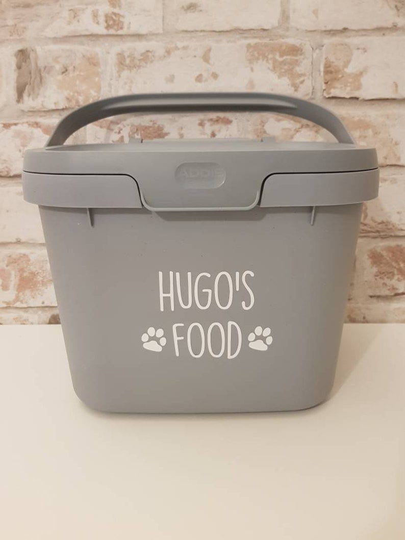 May include: A gray plastic container with a handle and a white label that says "Hugo's Food" with paw prints on either side of the word "Food".
