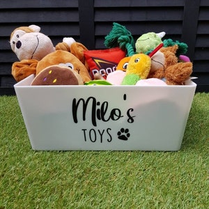 Op de afbeelding: Een witte plastic opbergbox met de woorden "Milo's Toys" en een pootprint op de voorkant. De box is gevuld met een verscheidenheid aan pluche speelgoed, waaronder een groen-wit pluche speelgoed, een bruin-wit pluche speelgoed en een rood-wit pluche speelgoed.