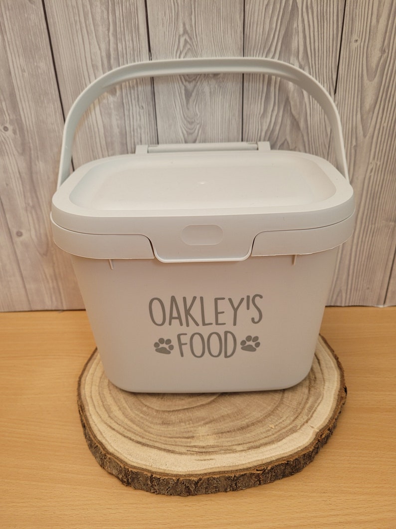 May include: A white plastic container with a handle and a lid. The container has the text "OAKLEY'S FOOD" with paw prints on either side of the word "FOOD".