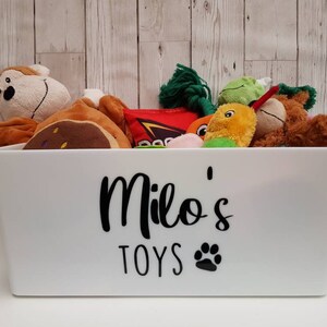 Op de afbeelding: Een witte plastic opbergbak met de tekst "Milo's Toys" en een pootprint. De bak is gevuld met een verscheidenheid aan knuffels, waaronder een aap, een bruine beer, een gele vogel en een groen-rood touwspeelgoed.