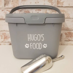May include: A gray plastic pet food container with a handle and a scoop. The container has the text "Hugo's Food" with paw prints on the front.