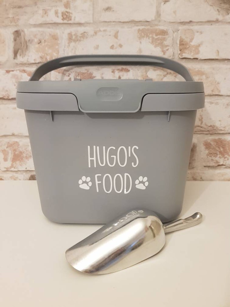 May include: A gray plastic pet food container with a handle and a scoop. The container has the text "Hugo's Food" with paw prints on the front.