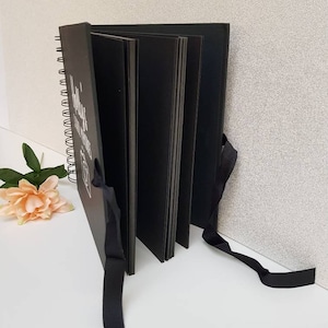 May include: Black spiral bound photo album with black ribbon ties. The cover has white text that reads "Hello, you are amazing".