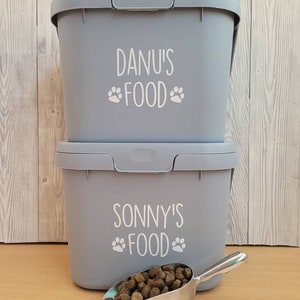 May include: Two gray plastic containers with white labels. The top container says "Danu's Food" with paw prints. The bottom container says "Sonny's Food" with paw prints. A silver scoop with dog food is in front of the containers.