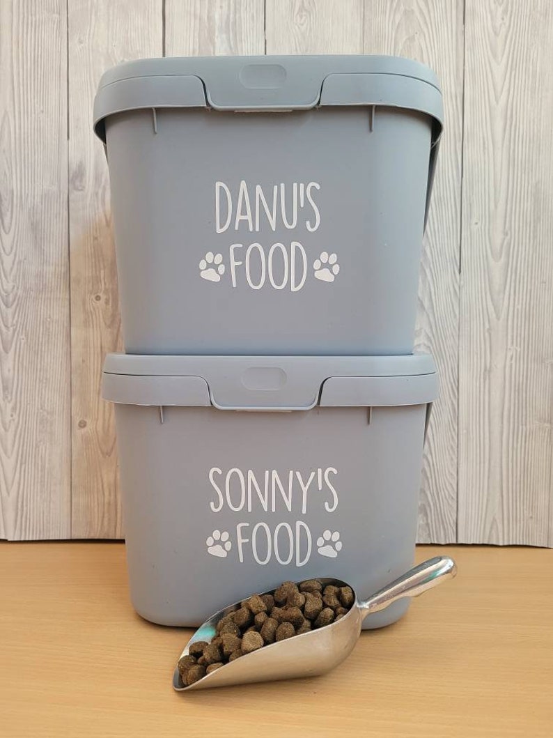 May include: Two gray plastic containers with white labels. The top container says "Danu's Food" with paw prints. The bottom container says "Sonny's Food" with paw prints. A silver scoop with dog food is in front of the containers.